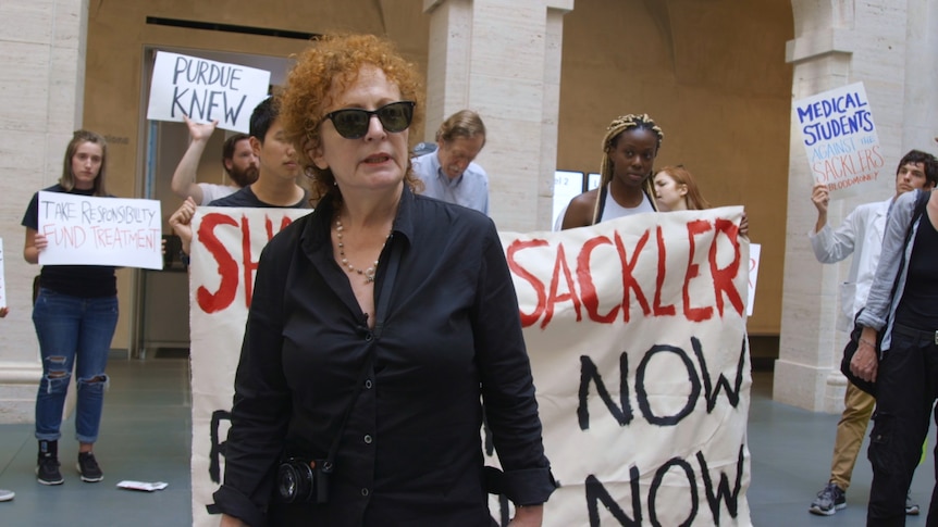 A red-haired woman in her late 60s, wearing sunglasses, stands in front of a protest outside an art gallery