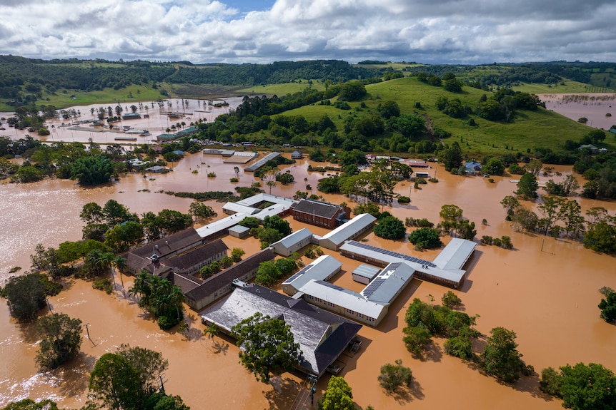 Aerial picture of a building complex inundated by muddy brown water, with inundated sheds and paddocks in the background.