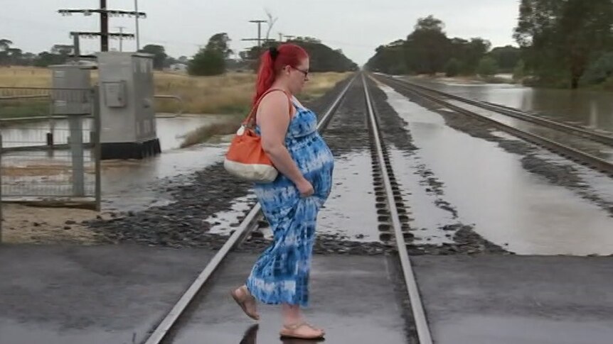 A pregnant woman walks across train tracks with floodwaters in the background.