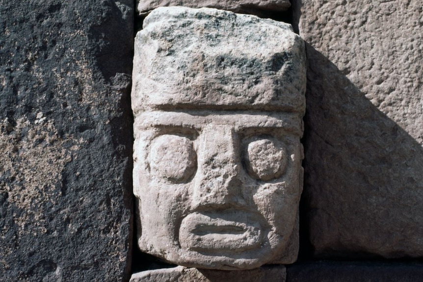 In a large block of rectangular stone, a face is carved: it has two large round eyes, a square-ish nose and a flat mouth.
