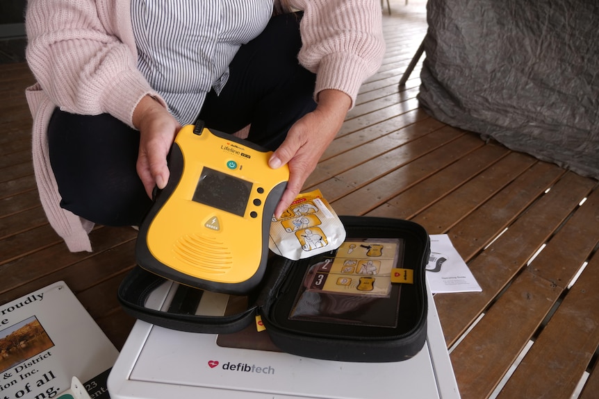 A pair of white female hands holds a yellow defibrillator out of a black case. It sits on a brown wooden deck. 