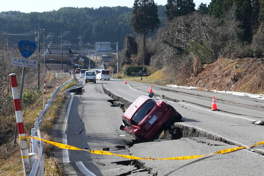 A red car is stuck between a large crack in the road after an earthquake.