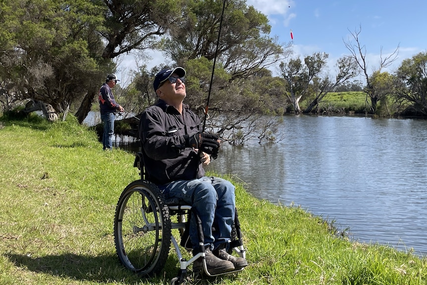 Man in wheel chair holding fishing rod next to river. 
