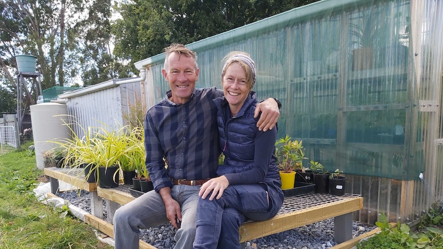 A man and a woman sit in front of a small greenhouse surrounded by young plants in pots