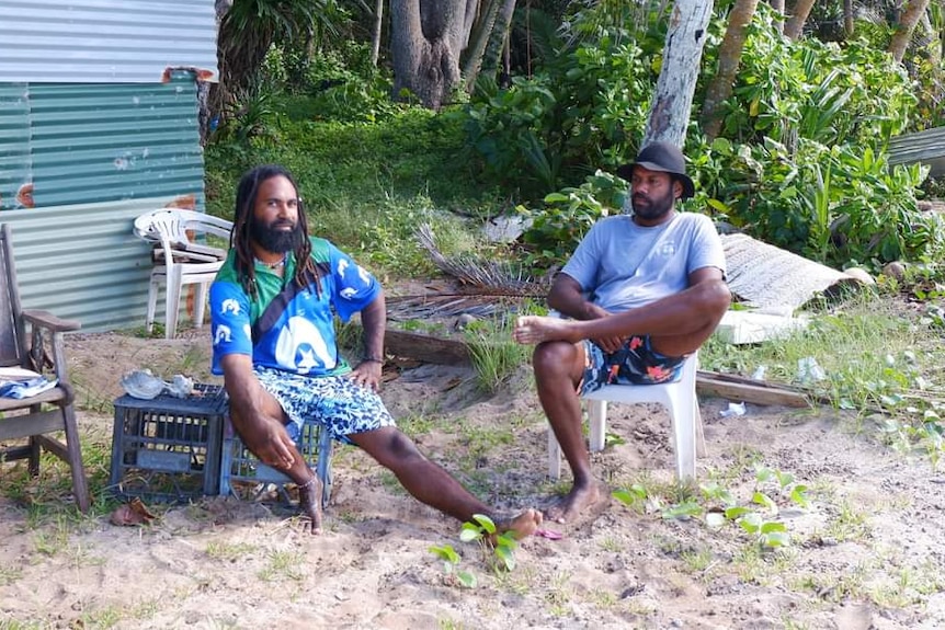Two men sitting in plastic chairs on the edge of a jungle on an island.
