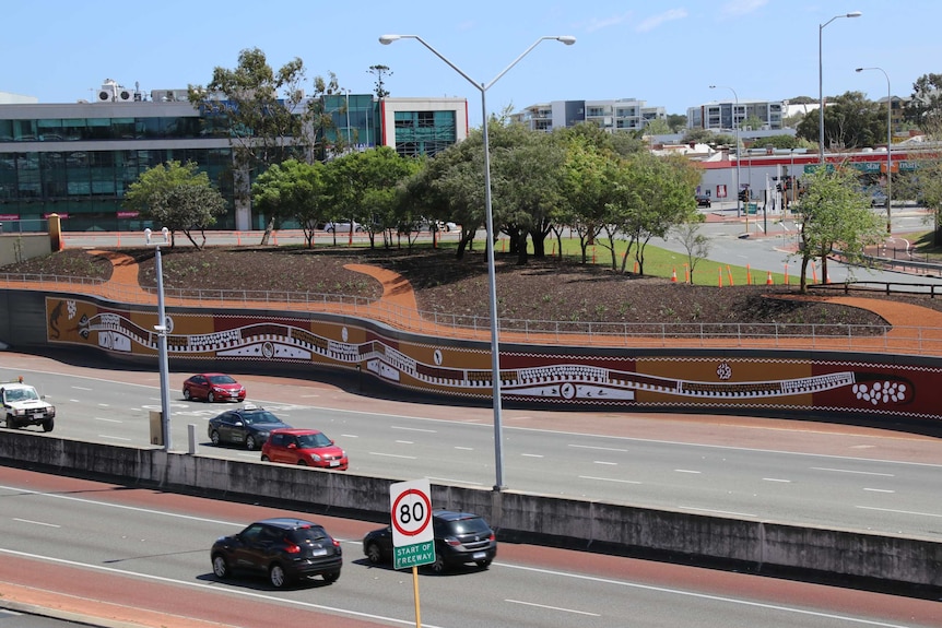 Aboriginal mural along wall of tunnel with cars passing in the foreground and buildings and trees in the background.