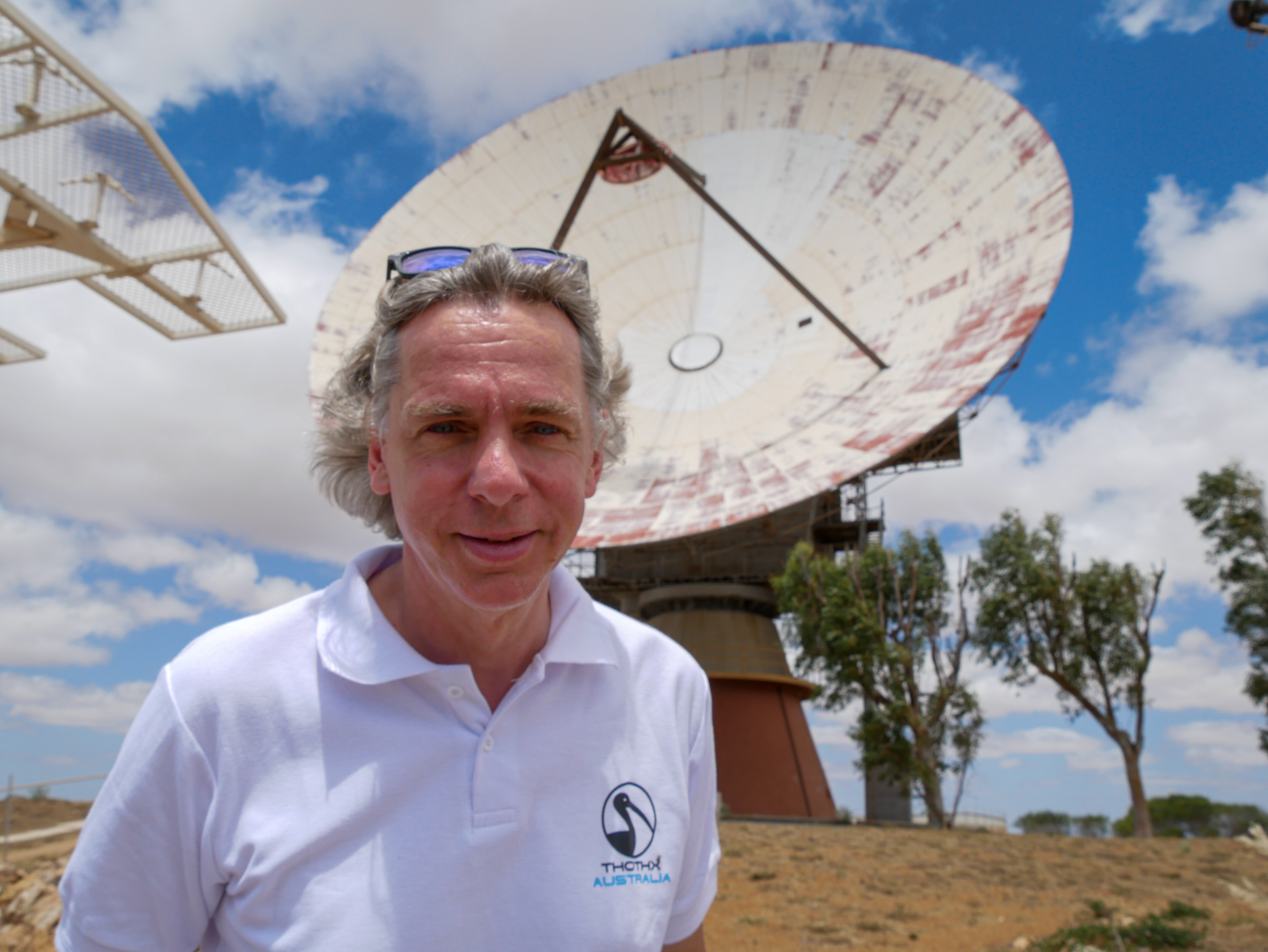 A middle aged man looks at the camera from in front of a huge satellite dish.