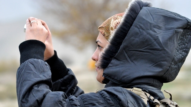 A woman in a hooded jacket looks through a camera, she is also wearing a scarf under the hood.