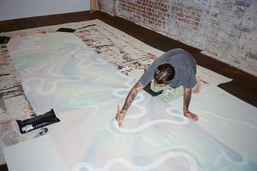 A man crouches down and paints pale swirling lines on a textile. 