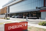 A picture of an emergency sign, with carpark, ambulances and windows in background, outside a hospital's Emergency Department 