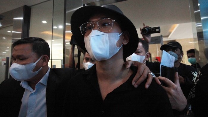 A bespectacled man wearing a black hat and face mask was surrounded by several reporters.