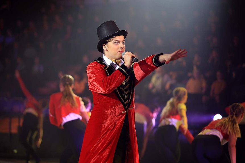 Blake Douglas wears a red coat and top hat while performing in a concert.