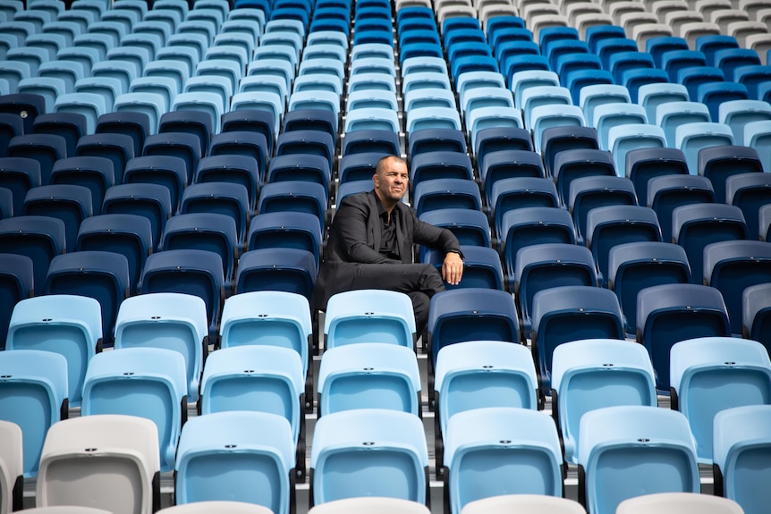 A man in a black suit and shirt sits alone in a stadium surrounded by empty blue chairs 