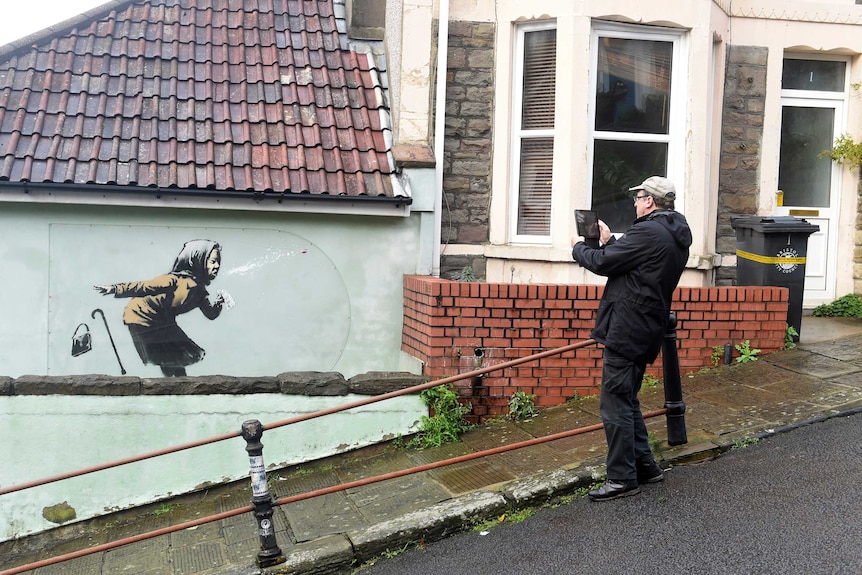 Un homme prend une photo sur une tablette en se tenant debout sur une route escarpée.  Il photographie un collage de street art d'une femme en train d'éternuer.