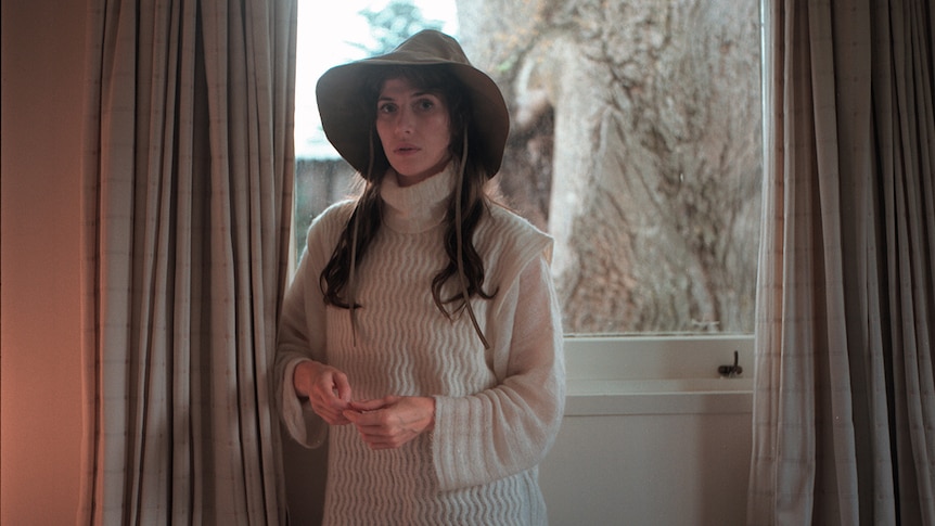 Aldous Harding stands before a window wearing a wide-brim hat and a white sweater