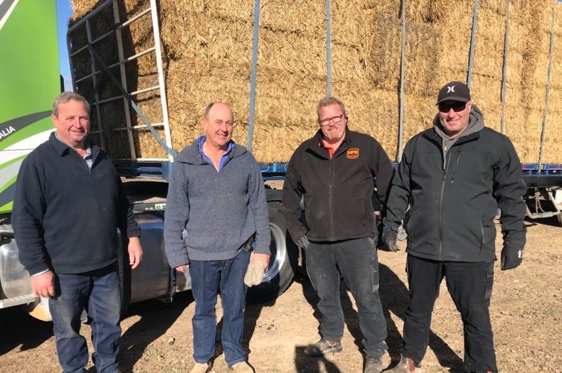 Four smiling men standing in front of a big truck filled with hay in a dry paddock.