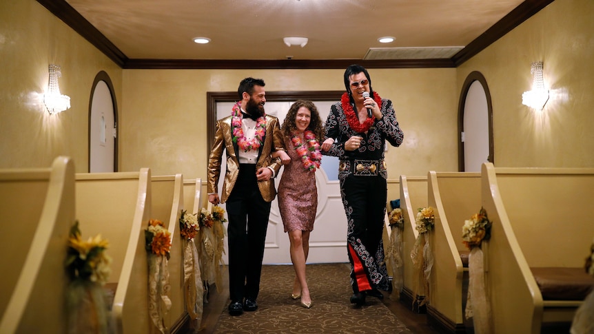a man and a woman with leis around their necks are walked down the aisle of a chapel by an Elvis Presley impersonator 
