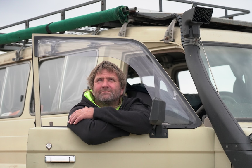 A man in a black jumper looks to the sky with his arms resting in the door of his beige car