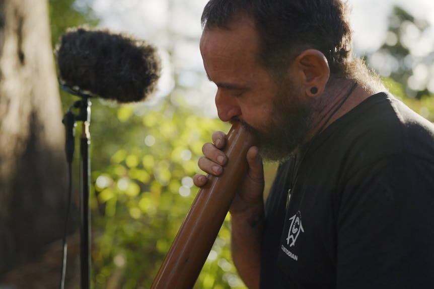 Man playing a didgeridoo as microphone records him.