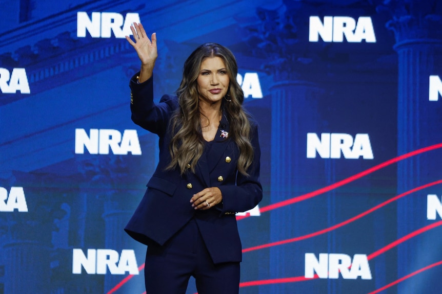 Kristi Noem looks glamorous standing and waving with the NRA logo behind her
