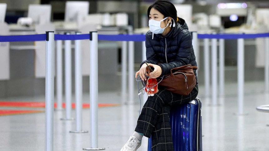 A young woman in a face mask sits on her suitcase at an airport