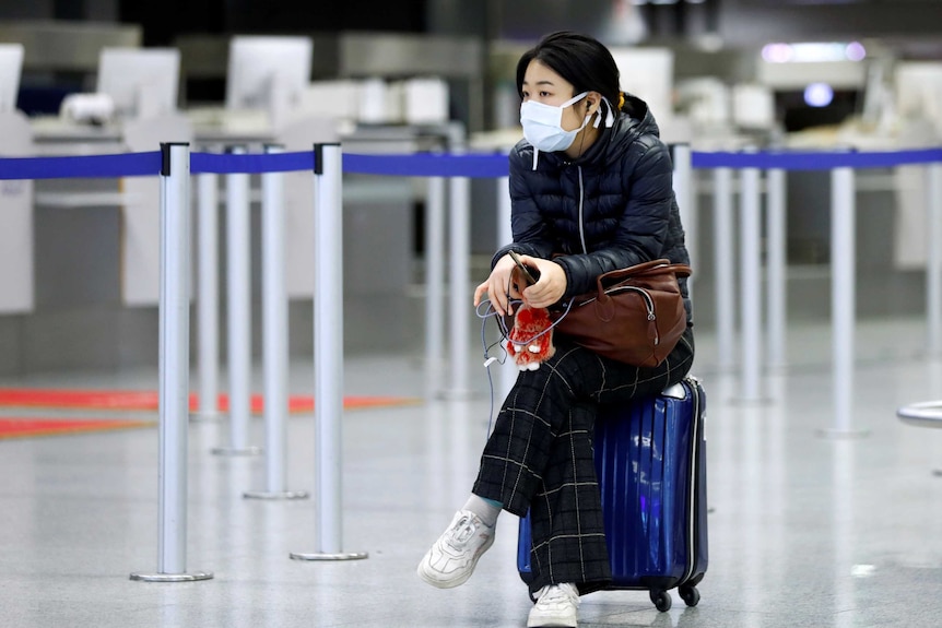 A young woman wearing a face mask sits on her suitcase in an airport