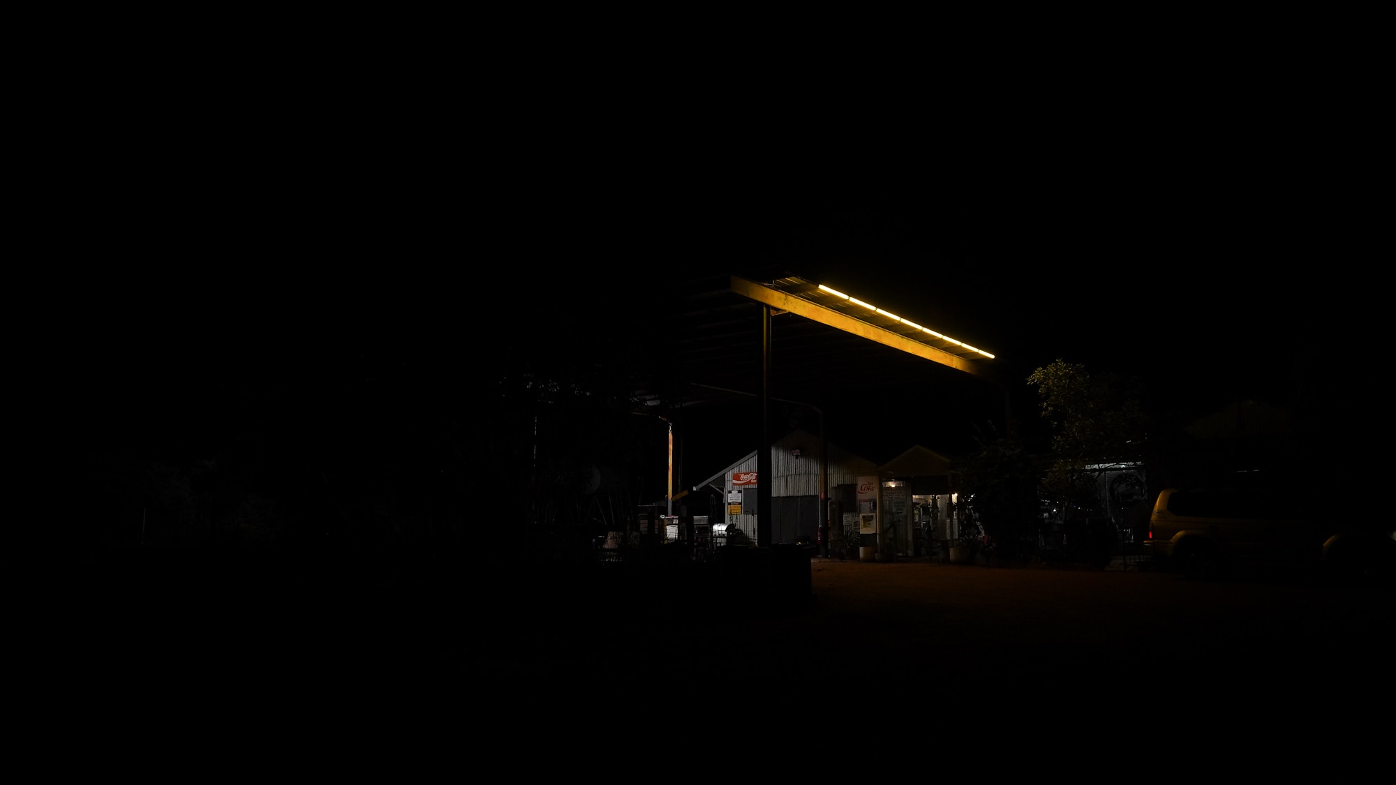 A dark image of the exterior of an outback tin shed hotel, with a yellow row of lights on the shelter roof offering dim light.