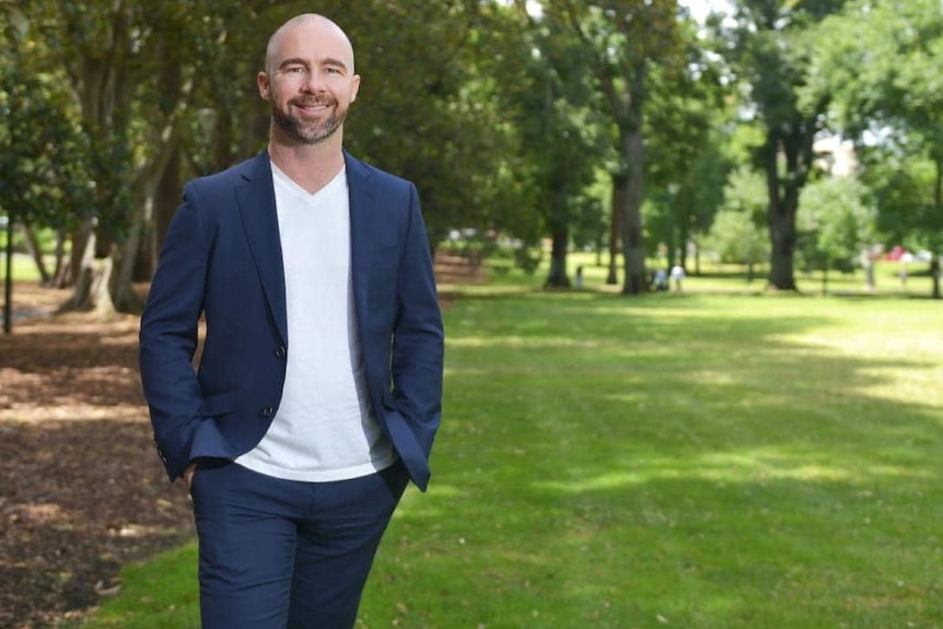 A bald man in a navy jacket and a white T-shirt standing in a park.