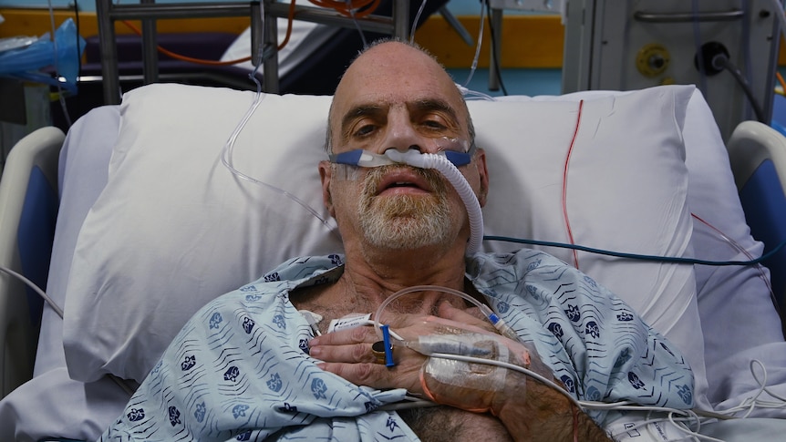 A COVID-19 patient has oxygen tubes in his nose and is hooked up to monitors in a hospital bed.