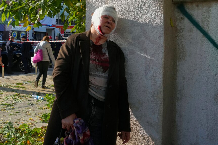 A middle-aged woman with a bandaged head and blood running from her chin leans against a wall.