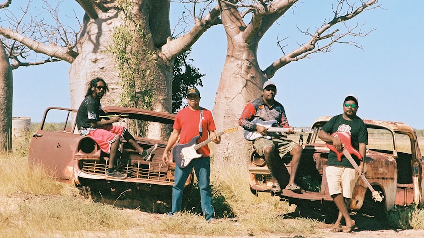 Four musicians holding guitars stand beside boab trees and rusty cars