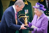 The Queen in a purple outfit smiles while receiving a gold trophy from her son, Prince Andrew