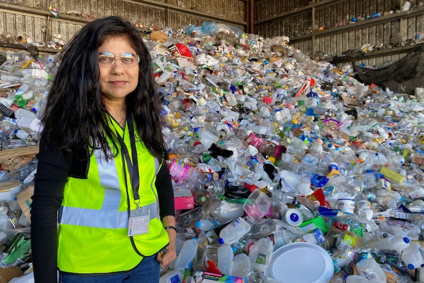 A woman with long dark hair stands in hi-vis vest at recycling plant in front of a large pile of plastic bottles