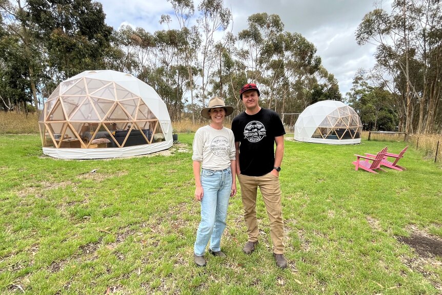 A man and a woman standing in front of two geodesic domes amid grass and trees
