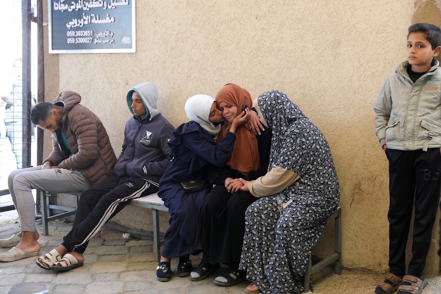 People sit on a bench waiting and a woman in a white headscarf hugs a woman in a tan headscaff