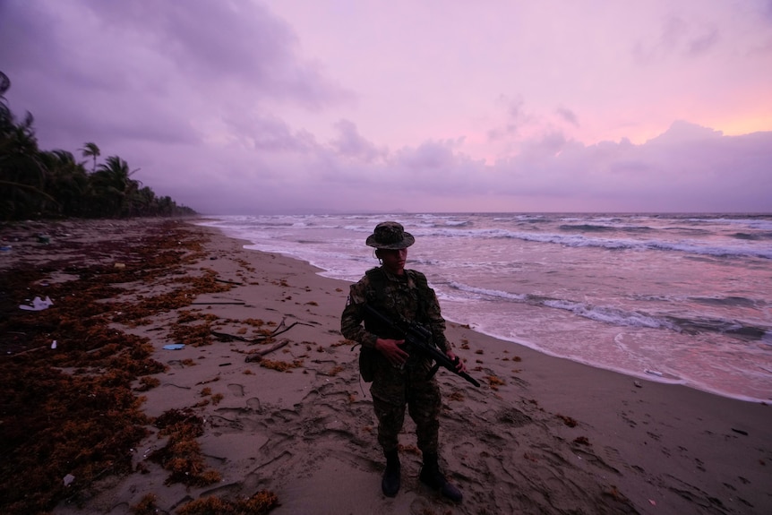 A Panamanian border officer stands in uniform with a rifle on a beach