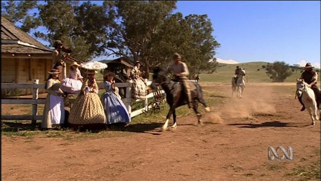 Horses gallop past a group of women in period costume near house