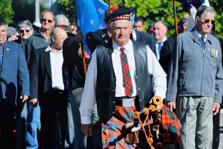 A man in a white shirt and blue and red tartan kilt holds bagpipes at a parade.
