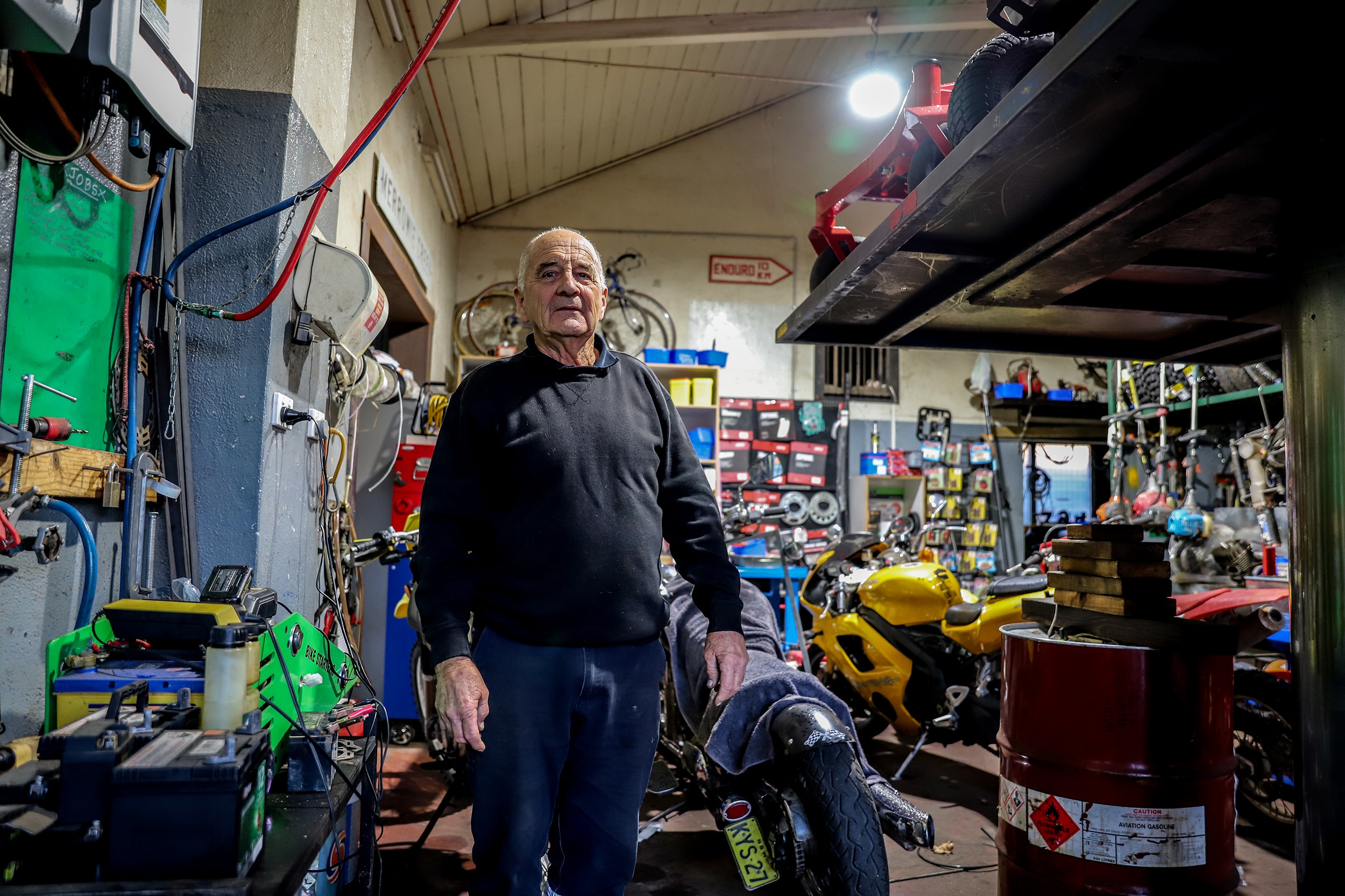 Older man wearing blue jumper stands in cluttered gar mechanic garage