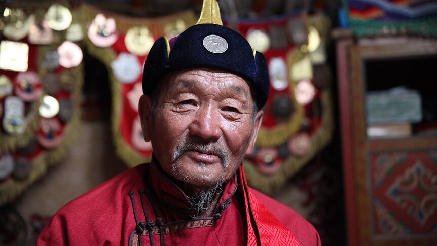 A Mongolian man in a hat and robe looks directly at the camera.