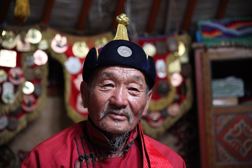 A Mongolian man in a hat and robe looks directly at the camera.