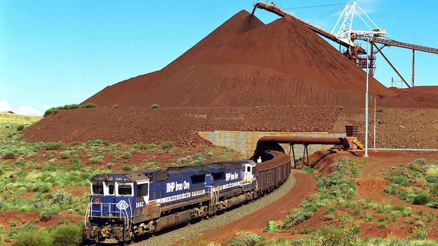 A blue train loaded with iron ore departs BHP's Yandi Mine in the Pilbara with a large dirt pile and loading belt behind.