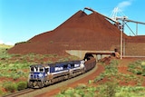 A blue train loaded with iron ore departs BHP's Yandi Mine in the Pilbara with a large dirt pile and loading belt behind.