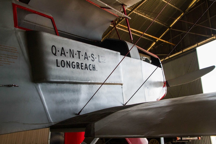 A close-up of a silver and red propeller plane in an old aviation hangar that says "Q.A.N.T.A.S Ltd Longreach" on the fuselage.