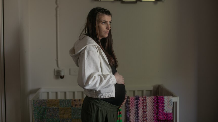 A young woman pregnant stands in front of a cot. She rests a hand on her stomach and looks forward.
