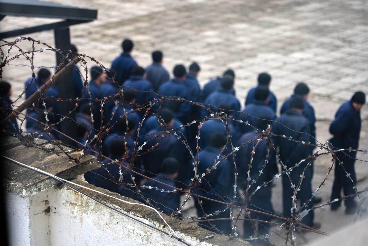 A group of prisoners wearing blue uniforms, seen from behind through a barbed-wire fence.