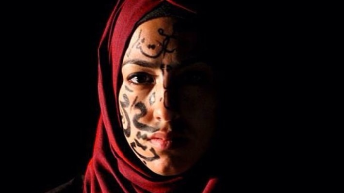 A woman wearing a red hijab with arabic letters on one side of her face, the other in darkness.