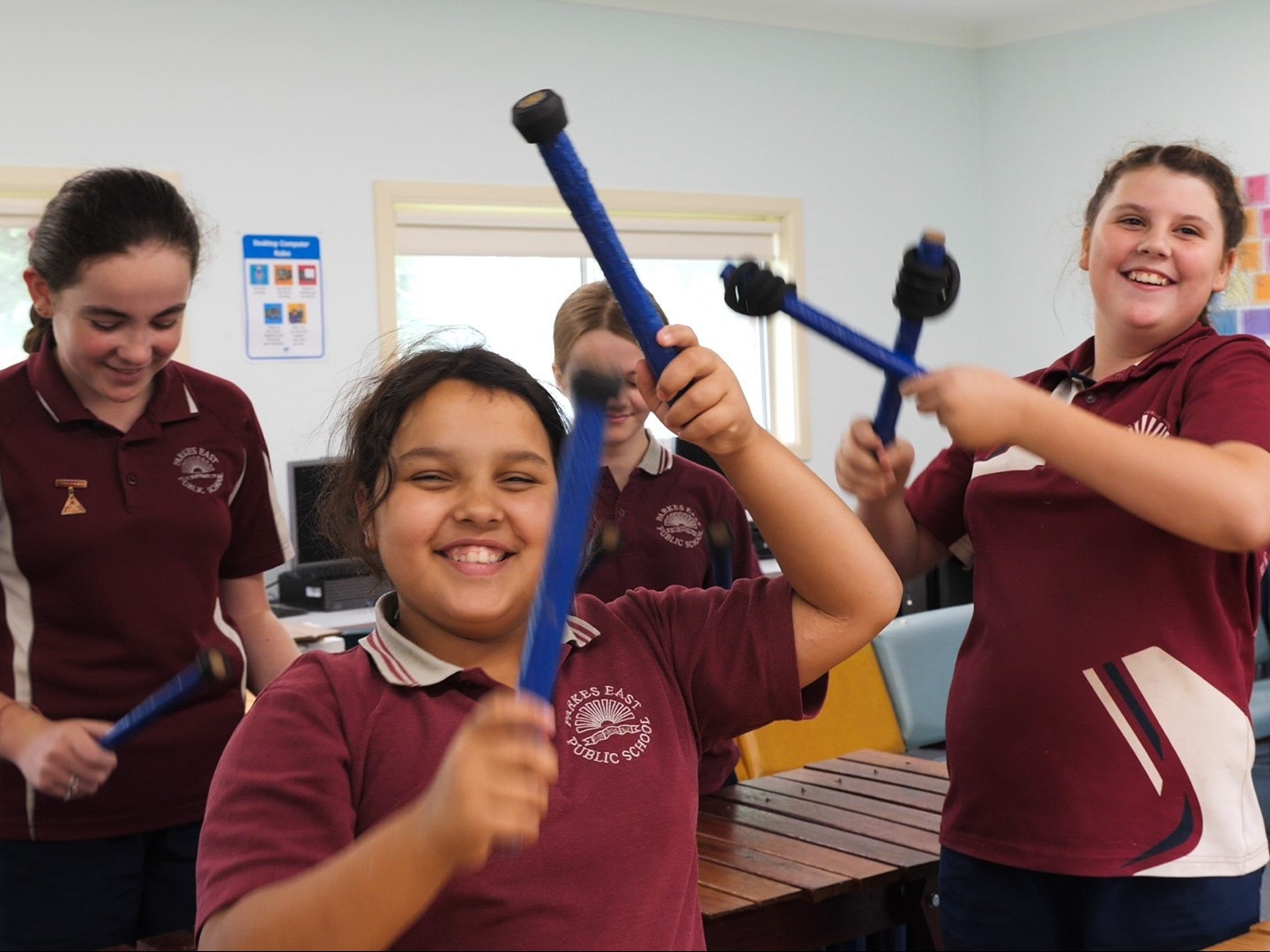 Four smiling girls in maroon school uniforms hold blue sticks above a wooden xylophone.