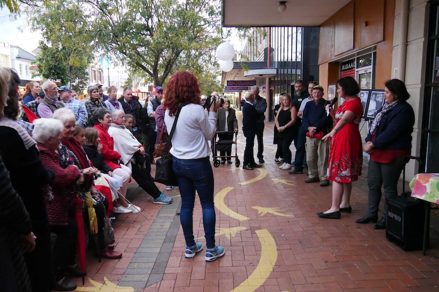 A crowd of people listening to a speaker on the footpath, in a main street of a country town.