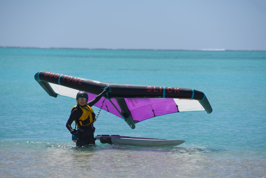 Helena Sahm stands in the water and holds a wing above a board. She's wearing a wetsuit and sunglasses.